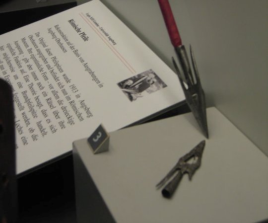 Een gesmede experimentele pijlpunt ligt in een vitrine naast een informatieblad.